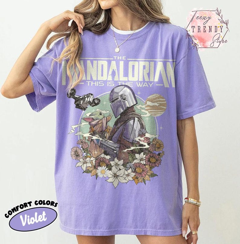 May include: A violet Comfort Colors t-shirt with a graphic of The Mandalorian and Grogu, surrounded by flowers. The design includes the text "THE MANDALORIAN - THIS IS THE WAY". The shirt has a relaxed fit.