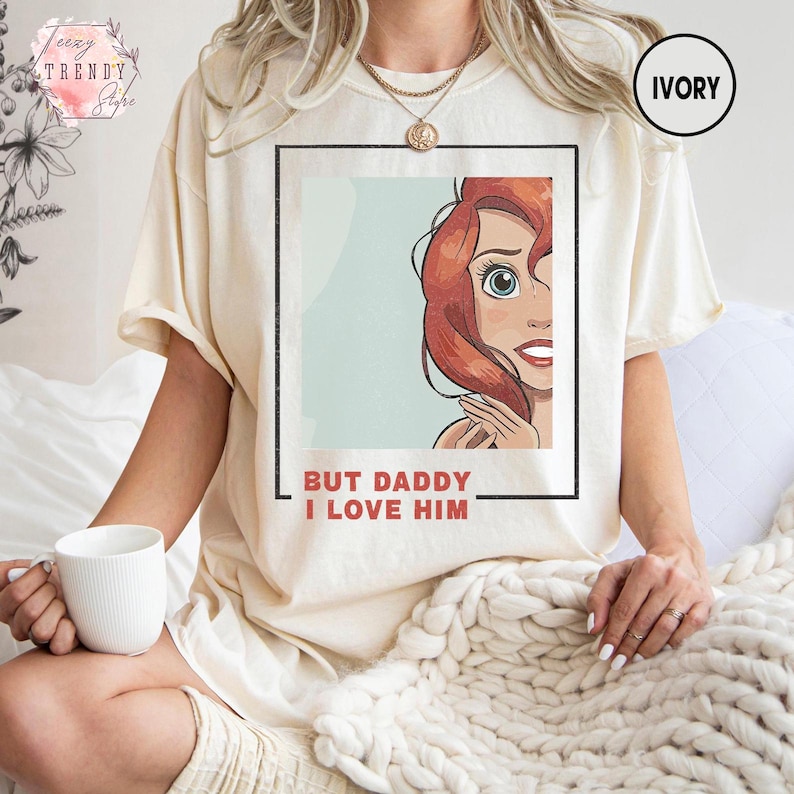 Puede incluir: Camiseta color marfil con un gr&aacute;fico de un personaje de dibujos animados con cabello rojo y el texto "BUT DADDY I LOVE HIM" en un marco negro. La camiseta tiene mangas cortas y un corte holgado. Parte de la colecci&oacute;n "Trendy Store".