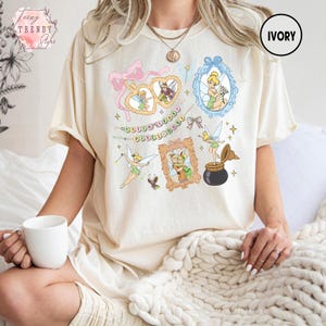 May include: Ivory-colored t-shirt featuring a whimsical design of Tinkerbell and other fairies in various frames and poses. The design includes pink bows, stars, and the text "Doodle Doodle Doodle".