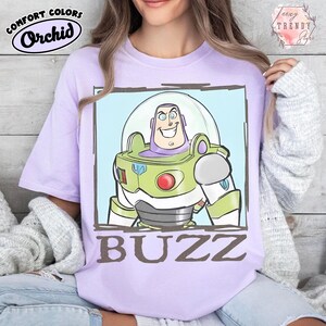 May include: A lilac t-shirt featuring a cartoon image of Buzz Lightyear in a green and white spacesuit. The character has a clear helmet and is set against a light blue background. The word "BUZZ" is printed in brown.