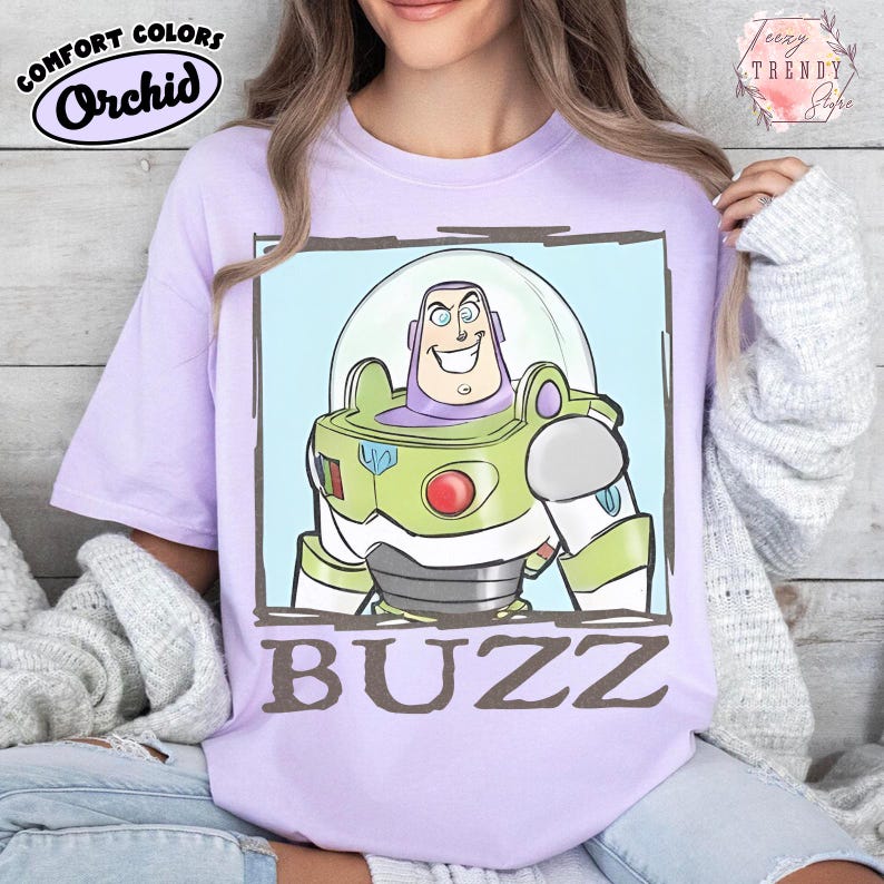 May include: A lilac t-shirt featuring a cartoon image of Buzz Lightyear in a green and white spacesuit. The character has a clear helmet and is set against a light blue background. The word "BUZZ" is printed in brown.