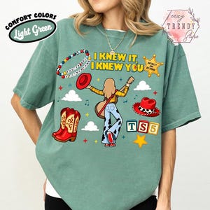 May include: A light green t-shirt featuring a graphic print of a person with long hair holding a guitar, surrounded by cowboy boots, a red hat, a sheriff badge with the text "INFINITY AND BEYOND", and blocks spelling "TS5". The text "I KNEW IT, I KNEW YOU" is printed above the figure.