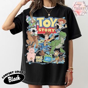 May include: A black short-sleeve t-shirt featuring a large graphic print of Toy Story characters including Woody, Buzz Lightyear, Jessie, Rex, Hamm, Bullseye, Slinky Dog, and an Alien. The text reads Toy Story, Since 1995, and Hi on a green square.