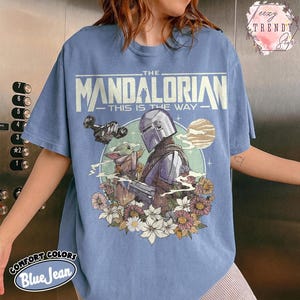 May include: A blue jean-coloured t-shirt with a graphic of The Mandalorian and Grogu surrounded by flowers. The text "THE MANDALORIAN - THIS IS THE WAY" is printed across the top. The shirt is made of Comfort Colors material.