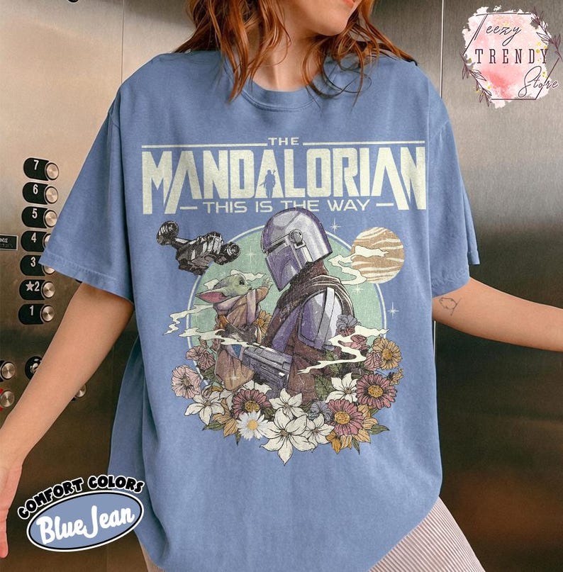 May include: A blue jean-coloured t-shirt with a graphic of The Mandalorian and Grogu surrounded by flowers. The text "THE MANDALORIAN - THIS IS THE WAY" is printed across the top. The shirt is made of Comfort Colors material.
