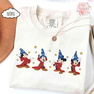 May include: Ivory-colored t-shirt featuring a graphic of Mickey Mouse in a wizard costume. The design shows Mickey in a red robe and blue wizard hat with stars. The shirt has a Comfort Colors tag.
