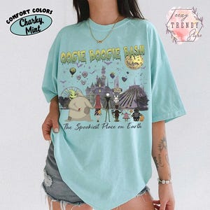 May include: A light mint green t-shirt featuring a graphic print with the text Oogie Boogie Bash and The Spookiest Place on Earth. The design includes characters from The Nightmare Before Christmas, a castle, a moon, and various park attractions.