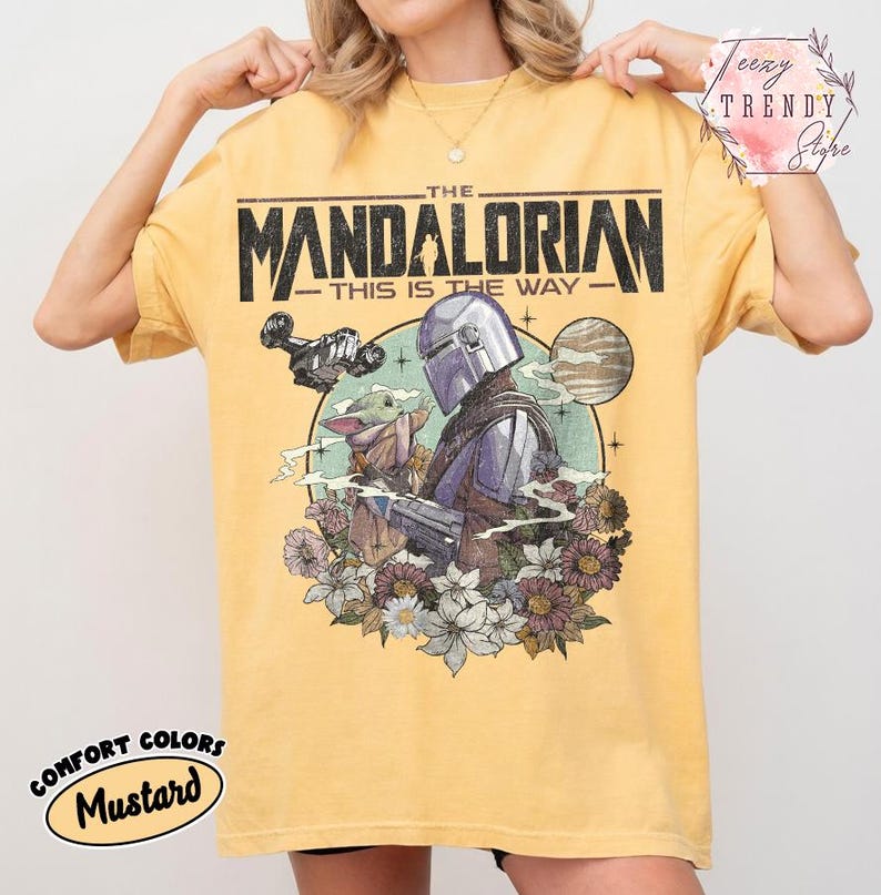May include: Mustard yellow t-shirt with a graphic from The Mandalorian. The design features the text "THE MANDALORIAN - THIS IS THE WAY" above an illustration of the Mandalorian, Grogu, and flowers. The shirt also has the words "COMFORT COLORS" and "Mustard" printed on it.