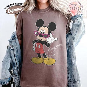 May include: Espresso-colored t-shirt featuring a vintage Mickey Mouse graphic wearing pink sunglasses. The cartoon character is in a confident pose, with the words "Mickey Mouse" in script. The shirt has a relaxed fit and is part of the "Comfort Colors" collection.