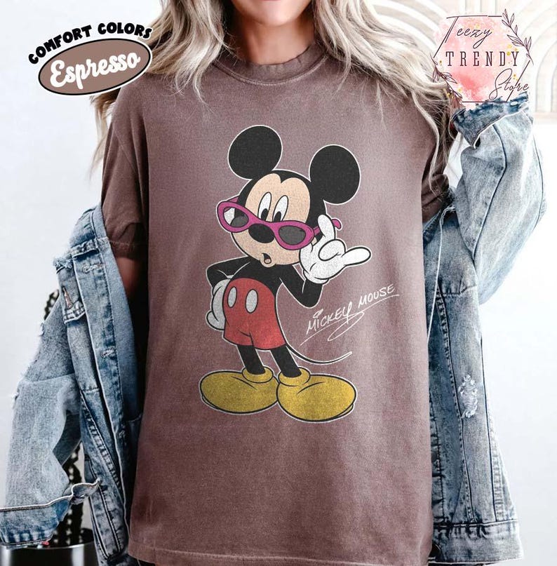 May include: Espresso-colored t-shirt featuring a vintage Mickey Mouse graphic wearing pink sunglasses. The cartoon character is in a confident pose, with the words "Mickey Mouse" in script. The shirt has a relaxed fit and is part of the "Comfort Colors" collection.