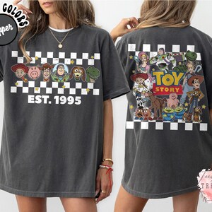Op de afbeelding: Donkergrijze t-shirt met personages uit de animatiefilm Toy Story. De voorkant van de shirt toont een dambordpatroon met de tekst "EST. 1995". De achterkant van de shirt heeft een grotere afbeelding van de Toy Story-personages.