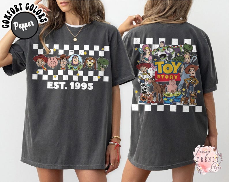 Op de afbeelding: Donkergrijze t-shirt met personages uit de animatiefilm Toy Story. De voorkant van de shirt toont een dambordpatroon met de tekst "EST. 1995". De achterkant van de shirt heeft een grotere afbeelding van de Toy Story-personages.