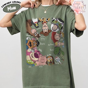 May include: Moss green t-shirt featuring a collage of Toy Story characters. The design includes Woody, Buzz Lightyear, and other characters from the animated film. The shirt has the words "COMFORT COLORS" and "Moss" printed on it.