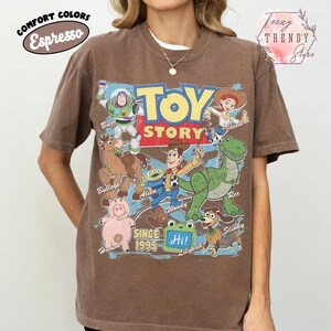 May include: An espresso brown short-sleeve t-shirt featuring a large graphic print of Toy Story characters including Buzz Lightyear, Jessie, Bullseye, Alien, Woody, Rex, Hamm, and Slinky. The text reads Toy Story, Since 1995, and Hi!.