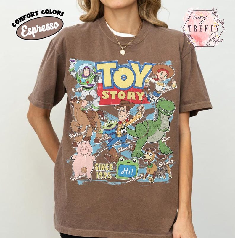 May include: An espresso brown short-sleeve t-shirt featuring a large graphic print of Toy Story characters including Buzz Lightyear, Jessie, Bullseye, Alien, Woody, Rex, Hamm, and Slinky. The text reads Toy Story, Since 1995, and Hi!.