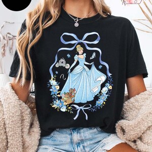 Peut inclure: T-shirt noir avec un motif Cendrillon. Le design comprend Cendrillon dans une robe bleue, un carrosse, un chausson de verre et des &eacute;l&eacute;ments floraux. Le mot "BLACK" est dans un cercle noir. Le t-shirt a un col rond.