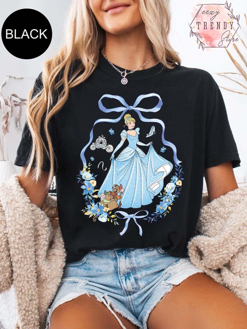 Peut inclure: T-shirt noir avec un motif Cendrillon. Le design comprend Cendrillon dans une robe bleue, un carrosse, un chausson de verre et des &eacute;l&eacute;ments floraux. Le mot "BLACK" est dans un cercle noir. Le t-shirt a un col rond.