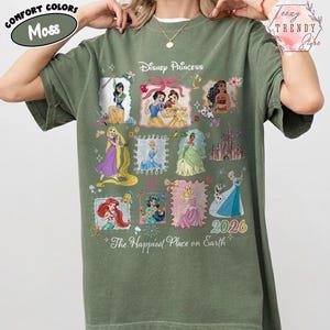 May include: A moss green t-shirt featuring Disney Princesses in framed illustrations. The shirt includes the text "Disney Princess" and "The Happiest Place on Earth". The year "2026" is also displayed.