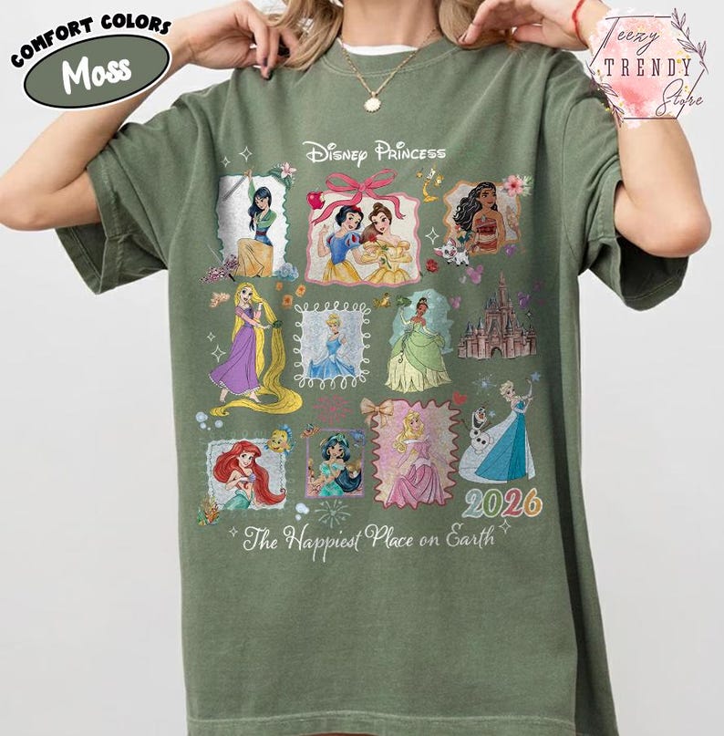 May include: A moss green t-shirt featuring Disney Princesses in framed illustrations. The shirt includes the text "Disney Princess" and "The Happiest Place on Earth". The year "2026" is also displayed.