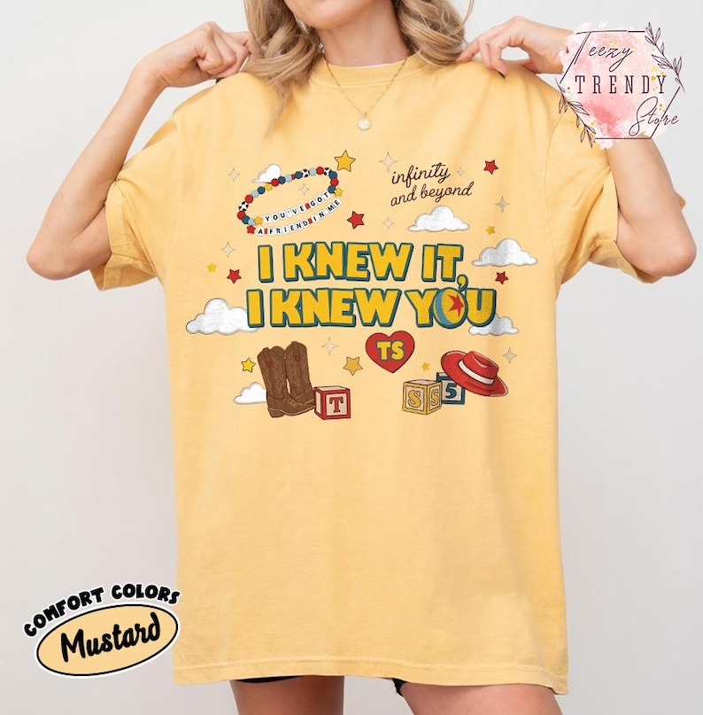 Puede incluir: Una camiseta color amarillo mostaza con un estampado gr&aacute;fico que incluye el texto I KNEW IT, I KNEW YOU, infinity and beyond, y YOU'VE GOT A FRIEND IN ME. El dise&ntilde;o presenta ilustraciones de botas vaqueras, un sombrero rojo, cubos y estrellas.
