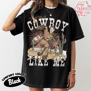 May include: A black short-sleeve t-shirt featuring a graphic print of a cartoon cowboy riding a horse in a desert landscape with cacti. The text reads You're a COWBOY LIKE ME. The design includes distressed white lettering and a sunburst background.