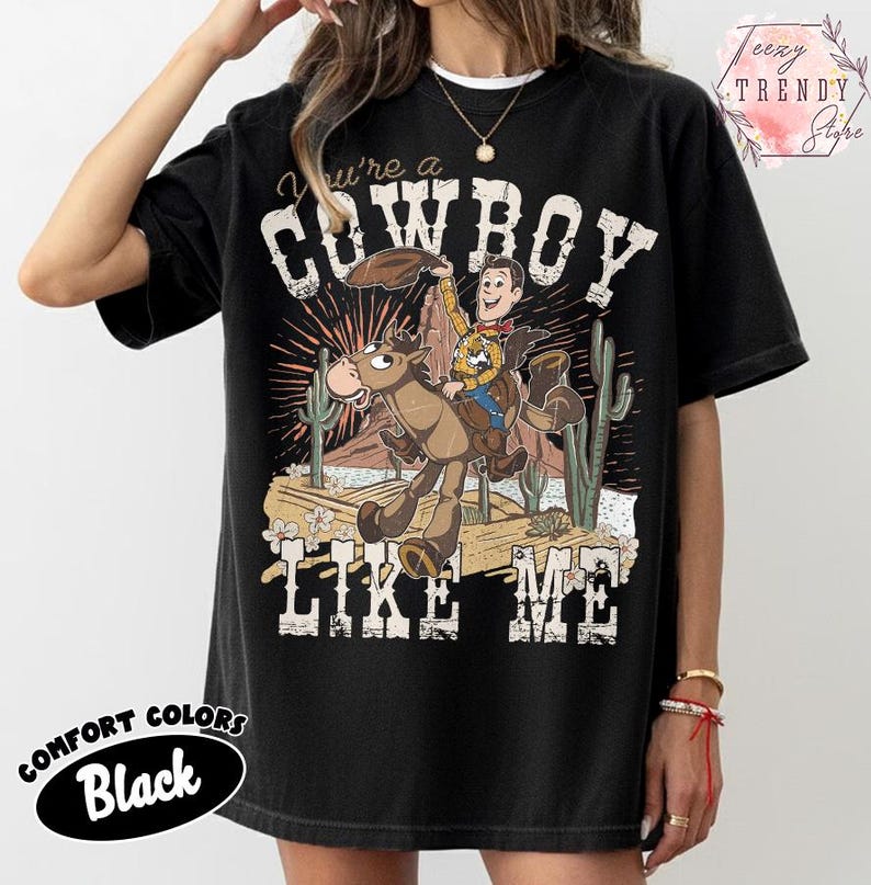 May include: A black short-sleeve t-shirt featuring a graphic print of a cartoon cowboy riding a horse in a desert landscape with cacti. The text reads You're a COWBOY LIKE ME. The design includes distressed white lettering and a sunburst background.