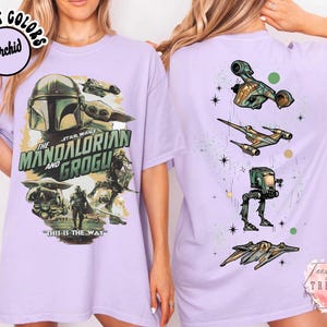 K&ouml;nnte beinhalten: Ein orchideenfarbenes T-Shirt mit einer Star Wars-Grafik auf der Vorderseite, die den Mandalorianer, Grogu sowie den Text "The Mandalorian and Grogu" und "This is the way" zeigt. Die R&uuml;ckseite zeigt vier Star Wars-Fahrzeuge untereinander.