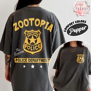 May include: Dark grey t-shirt featuring a yellow "Zootopia Police" badge design on the back and front. The back design includes the text "Zootopia Police Department" and "Now Recruiting!". The shirt is a comfort color.