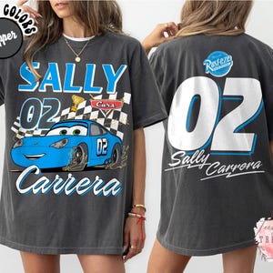 May include: Dark gray t-shirt featuring a blue cartoon car with the text "SALLY 02 Carrera" on the front. The back of the shirt displays a large "02" and "Sally Carrera" text. The shirt has a vintage, distressed look.