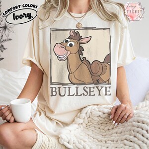 May include: A white ceramic mug with a black handle and the text "But First, Coffee" in a bold, black font. The mug is set against a blurred background. The mug appears to be approximately 10 cm tall.