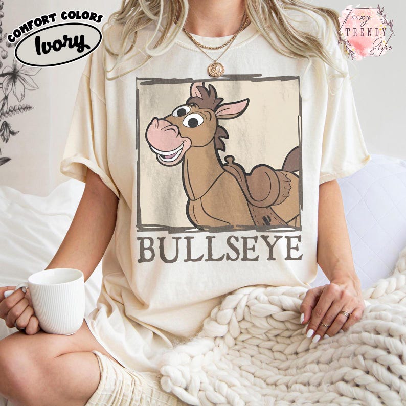 May include: A white ceramic mug with a black handle and the text "But First, Coffee" in a bold, black font. The mug is set against a blurred background. The mug appears to be approximately 10 cm tall.