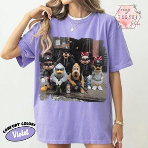 May include: A violet short-sleeved t-shirt featuring a graphic print of cartoon characters wearing sunglasses and baseball caps. The design shows Mickey Mouse, Goofy, Minnie Mouse, Donald Duck, Daisy Duck, and Pluto in an urban setting.