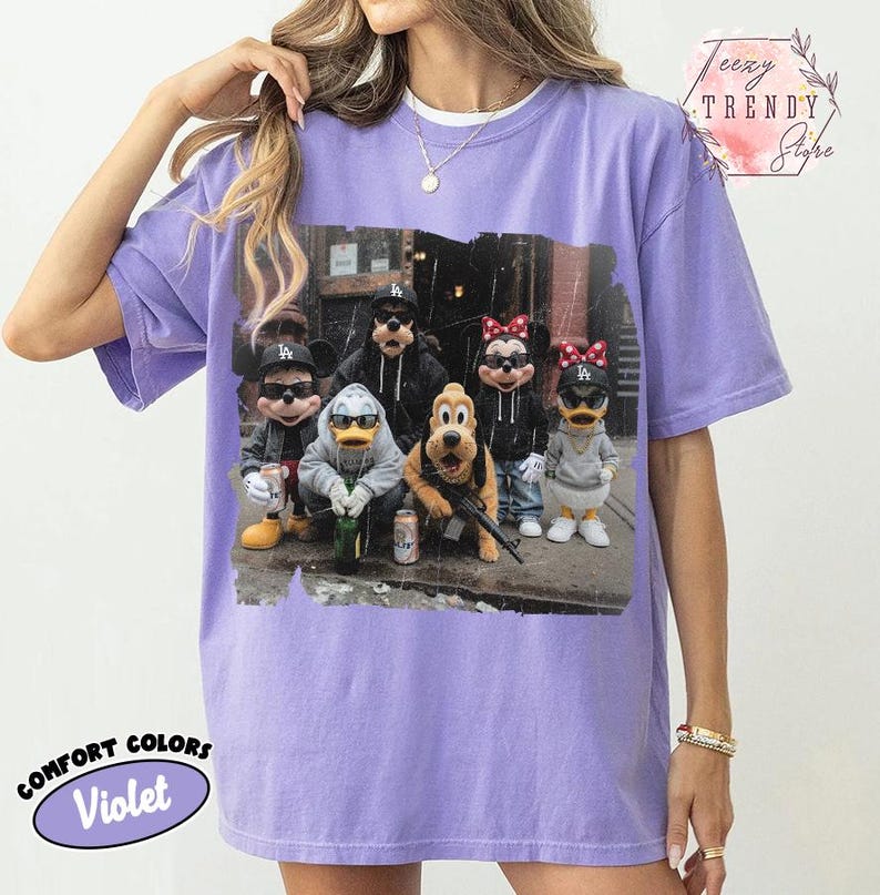 May include: A violet short-sleeved t-shirt featuring a graphic print of cartoon characters wearing sunglasses and baseball caps. The design shows Mickey Mouse, Goofy, Minnie Mouse, Donald Duck, Daisy Duck, and Pluto in an urban setting.