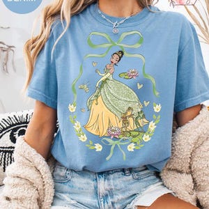Peut inclure: T-shirt bleu clair avec une illustration de dessin anim&eacute; d'une princesse en robe verte et jaune, entour&eacute;e d'une couronne florale et de grenouilles. Le t-shirt porte l'inscription "Washed Denim".