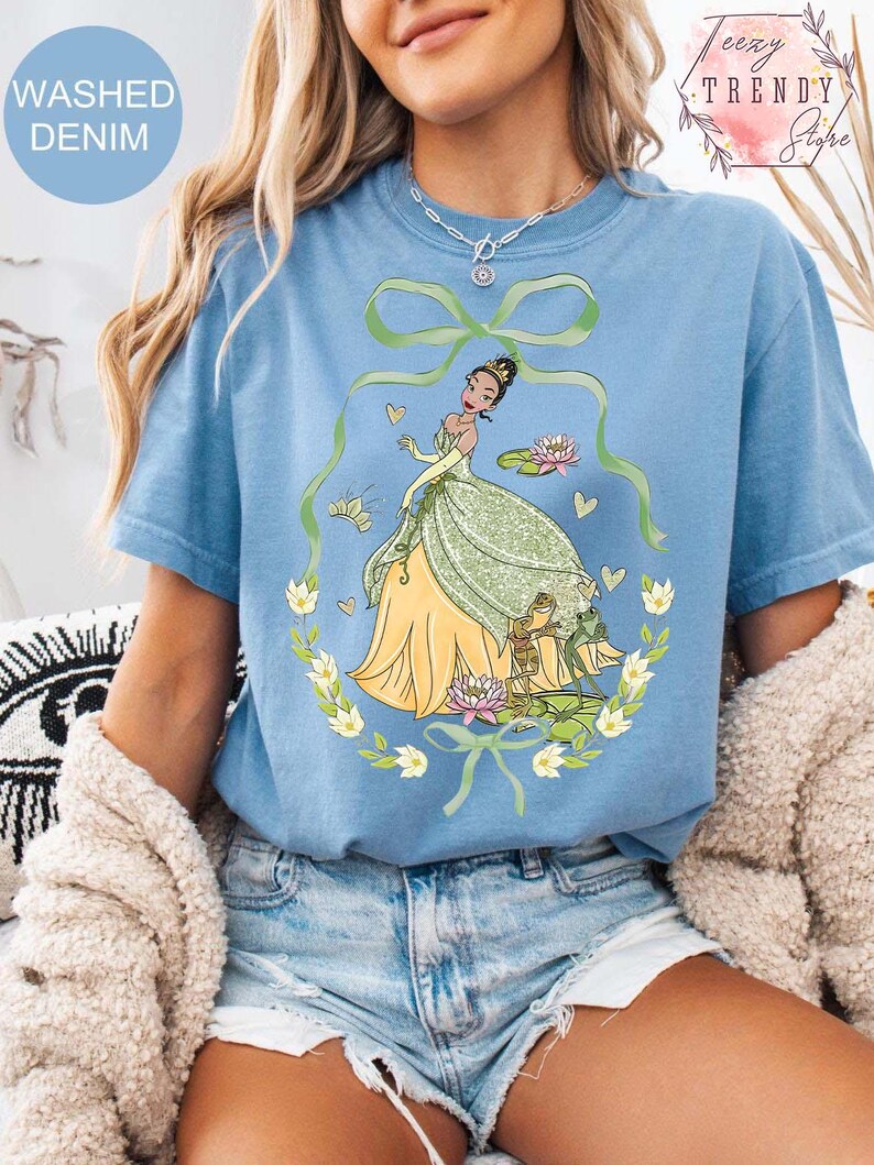Peut inclure: T-shirt bleu clair avec une illustration de dessin anim&eacute; d'une princesse en robe verte et jaune, entour&eacute;e d'une couronne florale et de grenouilles. Le t-shirt porte l'inscription "Washed Denim".