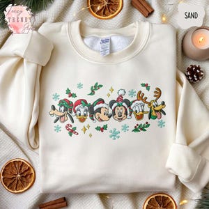 May include: Cream-colored sweatshirt featuring embroidered Disney characters in Christmas attire. The design includes Mickey Mouse, Minnie Mouse, Donald Duck, Goofy, and Pluto, with snowflakes and holly accents. The word "SAND" is visible in the top right corner.