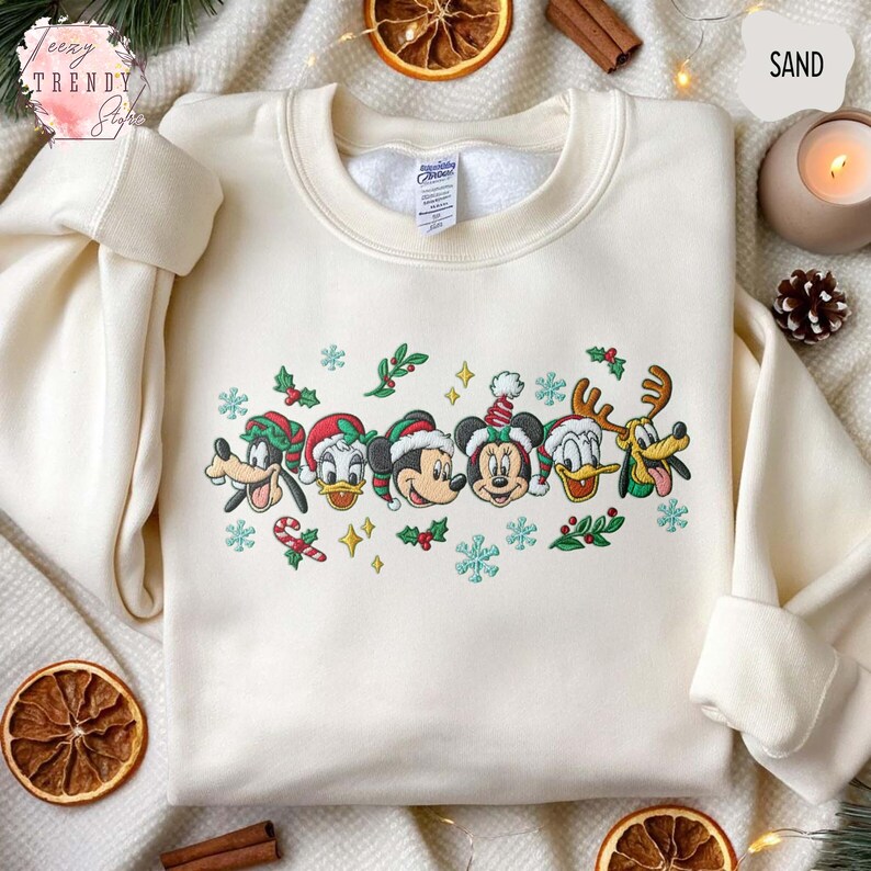 May include: Cream-colored sweatshirt featuring embroidered Disney characters in Christmas attire. The design includes Mickey Mouse, Minnie Mouse, Donald Duck, Goofy, and Pluto, with snowflakes and holly accents. The word "SAND" is visible in the top right corner.
