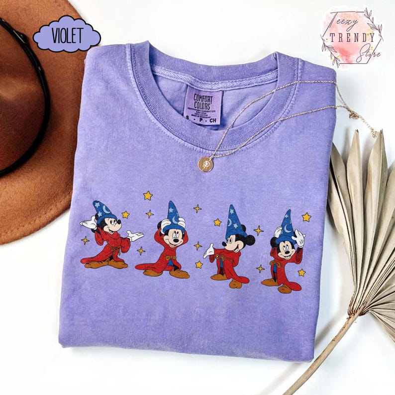 May include: Violet-colored t-shirt featuring a graphic of Mickey Mouse in a wizard costume with stars. The shirt has a crew neck and short sleeves. The text "Comfort Colors" is visible on the tag. A gold necklace is draped across the shirt.