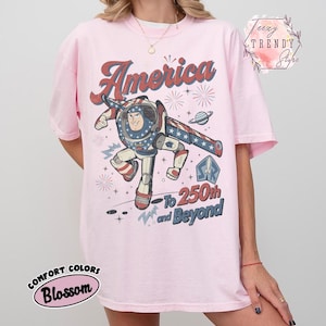 Puede incluir: Una camiseta rosa claro con un gr&aacute;fico de un personaje de guardi&aacute;n espacial con traje rojo, blanco y azul. El texto dice America, To 250th and Beyond, con ilustraciones de fuegos artificiales y un planeta. Camiseta de manga corta y cuello redondo.