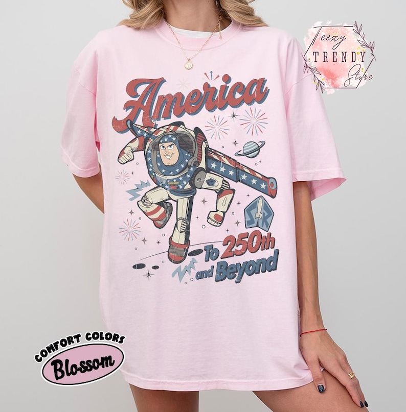 Puede incluir: Una camiseta rosa claro con un gr&aacute;fico de un personaje de guardi&aacute;n espacial con traje rojo, blanco y azul. El texto dice America, To 250th and Beyond, con ilustraciones de fuegos artificiales y un planeta. Camiseta de manga corta y cuello redondo.