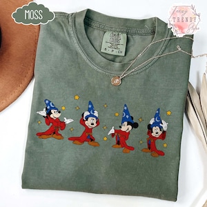May include: Moss green Comfort Colors t-shirt featuring a graphic of Mickey Mouse in a wizard costume. The design includes Mickey in a red robe and blue wizard hat with stars. A silver necklace with a pendant is also visible.