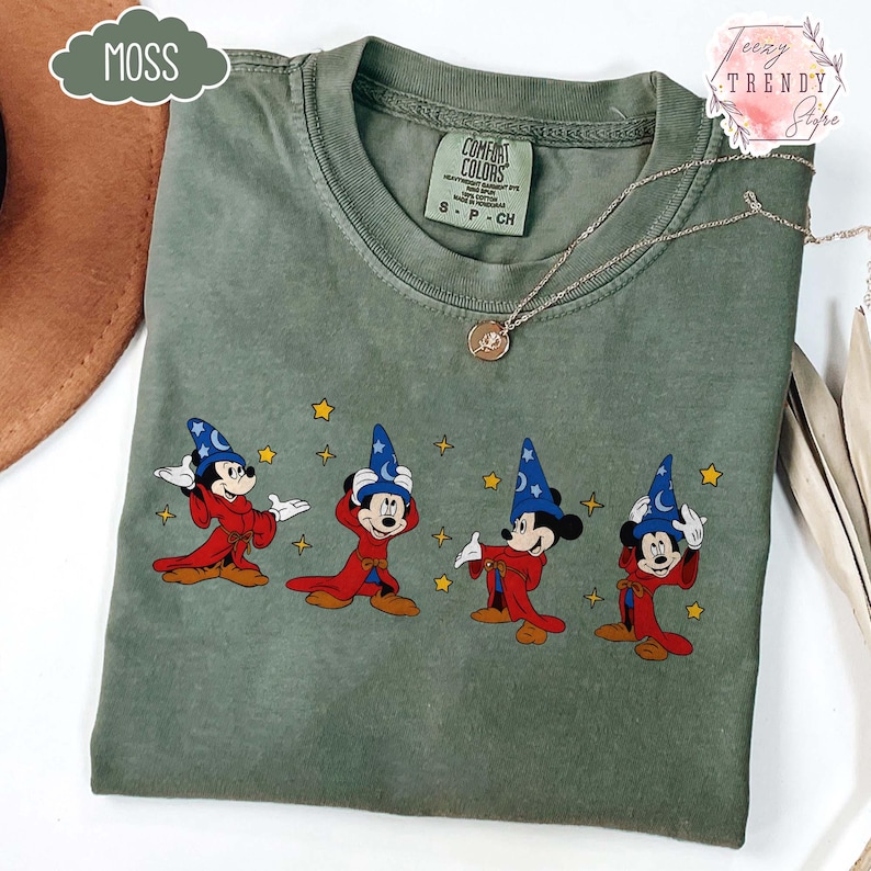 May include: Moss green Comfort Colors t-shirt featuring a graphic of Mickey Mouse in a wizard costume. The design includes Mickey in a red robe and blue wizard hat with stars. A silver necklace with a pendant is also visible.