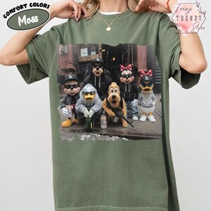 May include: A moss green short-sleeve t-shirt featuring a graphic print of five cartoon characters wearing black baseball caps and sunglasses. The characters are dressed in casual streetwear, including hoodies and jackets, and are posed together in an urban setting.