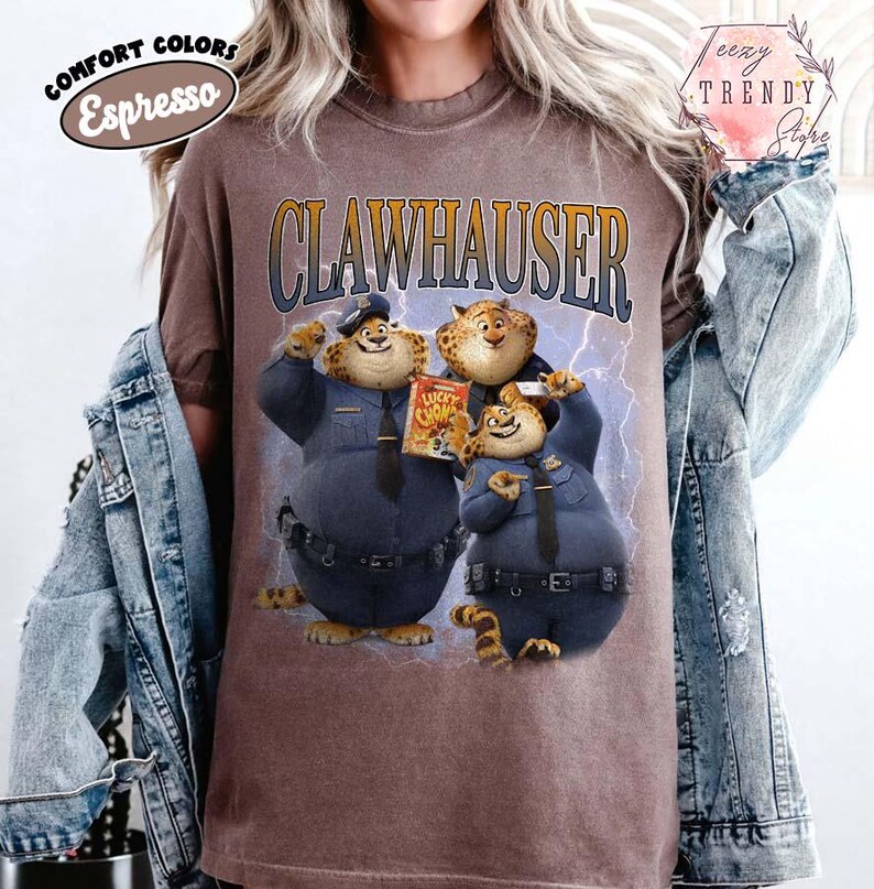 Puede incluir: Camiseta color espresso con la palabra "CLAWHAUSER" sobre un gr&aacute;fico de tres personajes de dibujos animados con uniformes de polic&iacute;a. Los personajes son como leopardos con manchas y sostienen una caja de cereales "Lucky Chows". La camiseta tiene las palabras "Comfort Colors".