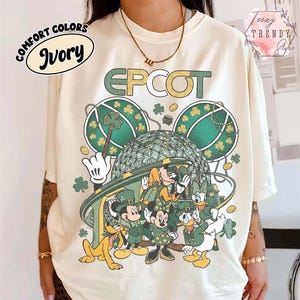 Puede incluir: Camiseta color marfil con un diseño del Día de San Patricio con la palabra "EPCOT" en verde. El gráfico incluye a Mickey Mouse, Minnie Mouse, Donald Duck y Pluto con trajes verdes, tréboles y un diseño verde y dorado.