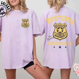 May include: Light purple t-shirt with a police badge graphic on the front and back. The front badge is gold and black, and the back has the word "ZOOTOPIA" in yellow. The shirt is a comfort colors style.