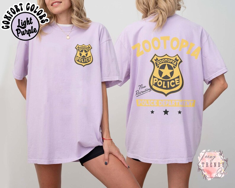 May include: Light purple t-shirt with a police badge graphic on the front and back. The front badge is gold and black, and the back has the word "ZOOTOPIA" in yellow. The shirt is a comfort colors style.