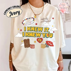 Puede incluir: Una camiseta color marfil con un estampado gr&aacute;fico que incluye el texto I KNEW IT, I KNEW YOU, infinity and beyond, y YOU'VE GOT A FRIEND IN ME. El dise&ntilde;o presenta ilustraciones de botas vaqueras marrones, un sombrero rojo, bloques y estrellas.