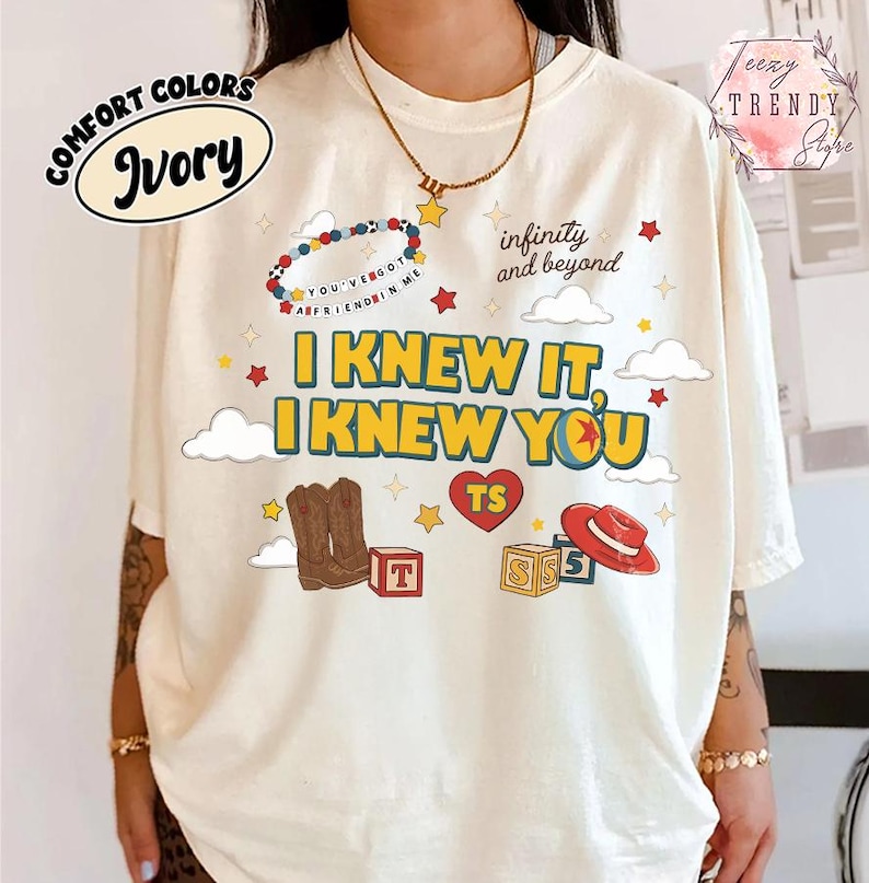 Puede incluir: Una camiseta color marfil con un estampado gr&aacute;fico que incluye el texto I KNEW IT, I KNEW YOU, infinity and beyond, y YOU'VE GOT A FRIEND IN ME. El dise&ntilde;o presenta ilustraciones de botas vaqueras marrones, un sombrero rojo, bloques y estrellas.