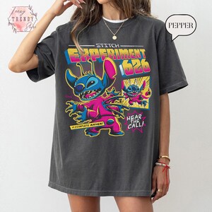 Pode incluir: T-shirt cinza escuro com um gr&aacute;fico colorido de Stitch de Lilo & Stitch. O design inclui o texto "Stitch Experiment 626" e "Hear the Call!" em estilo cartoon. A t-shirt tem um corte folgado.